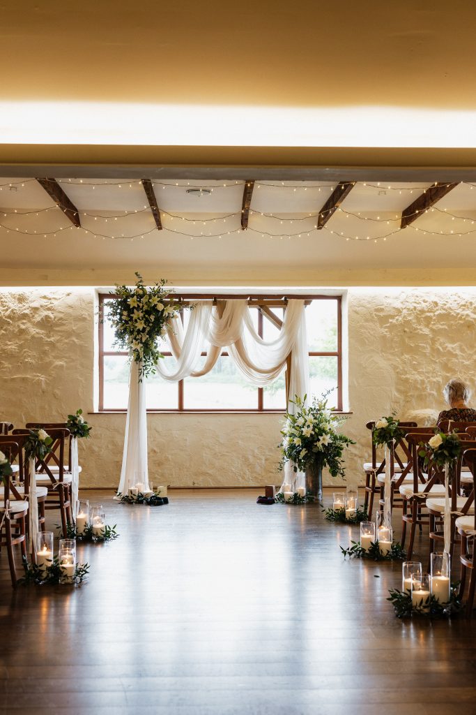 jo paul - fraoch floral design fraoch floral design wedding ceremony setup: central aisle with floral arrangements, sheer white drapery, and warm string lights in a rustic hall.