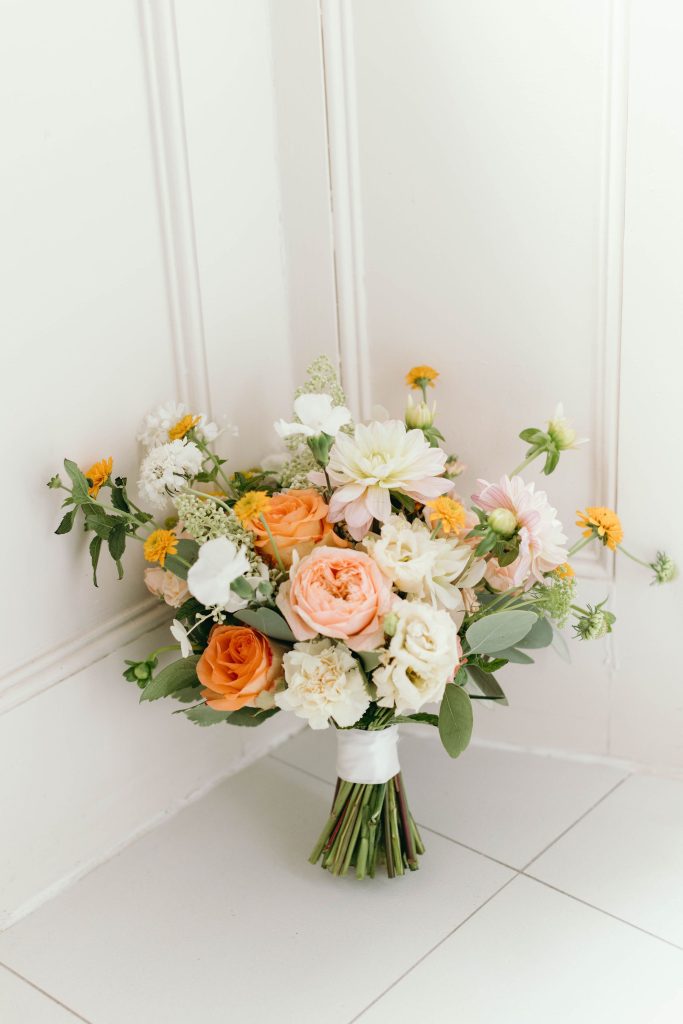 fraoch floral design bouquet of peach, white, and pink flowers with greenery, tied at the stems, resting on a white tiled floor against a pale wall.