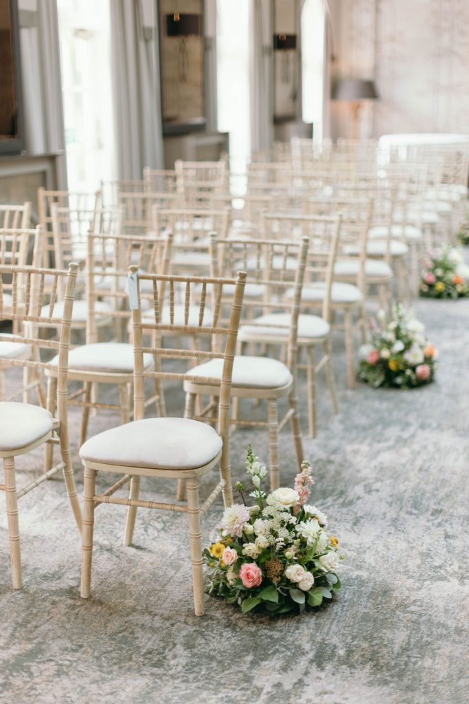 fraoch floral design rows of beige chiavari chairs set up for a ceremony with flower arrangements along the aisle on a gravel surface.