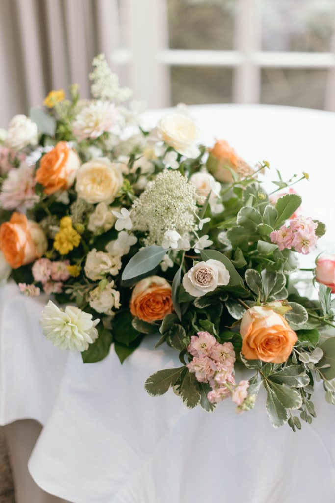 fraoch floral design bouquet of cream, peach, and pink flowers with greenery on a white table by a window (soft focus).