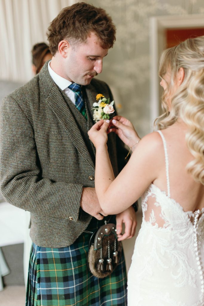 fraoch floral design bride adjusts a floral boutonniere on the groom's jacket as they prep for their wedding, with the groom in a tweed suit and tartan kilt nearby.