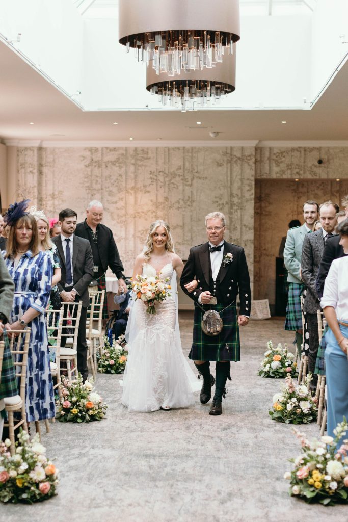 fraoch floral design bride in a lace wedding gown walks down the aisle with a man in a kilt, guests seated on either side and floral arrangements lining the carpeted aisle.