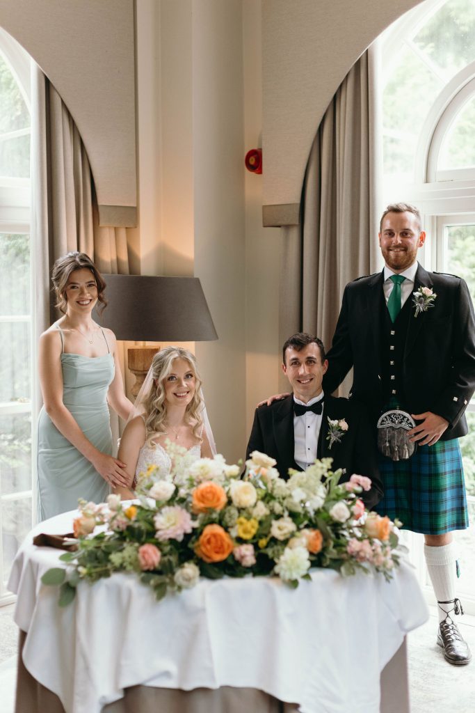 fraoch floral design bride and bridesmaids posing around a table with a lush flower centerpiece indoors, groomsmen standing behind them.