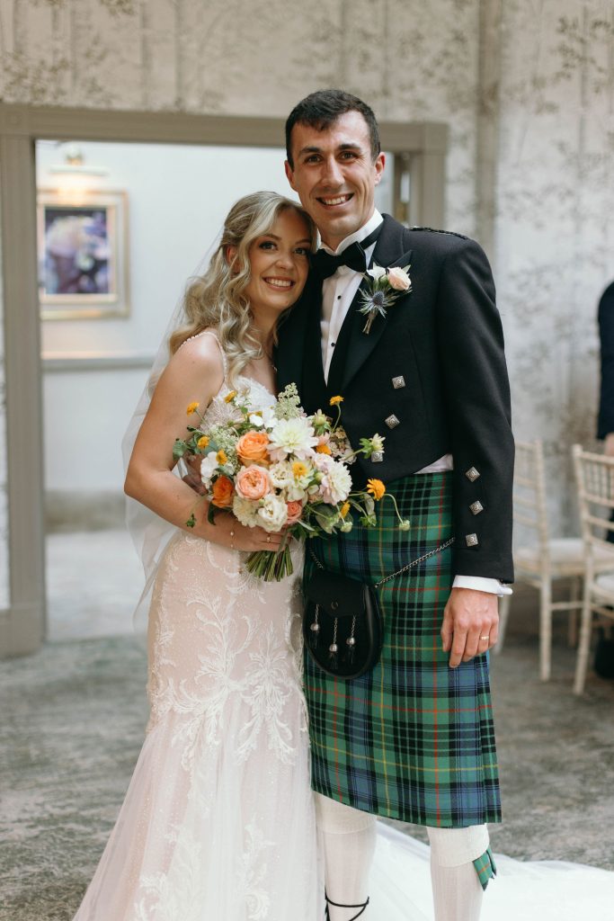fraoch floral design bride and groom smile for a wedding photo; bride in a white lace gown holds a pastel bouquet. groom wears a black jacket and green tartan kilt.