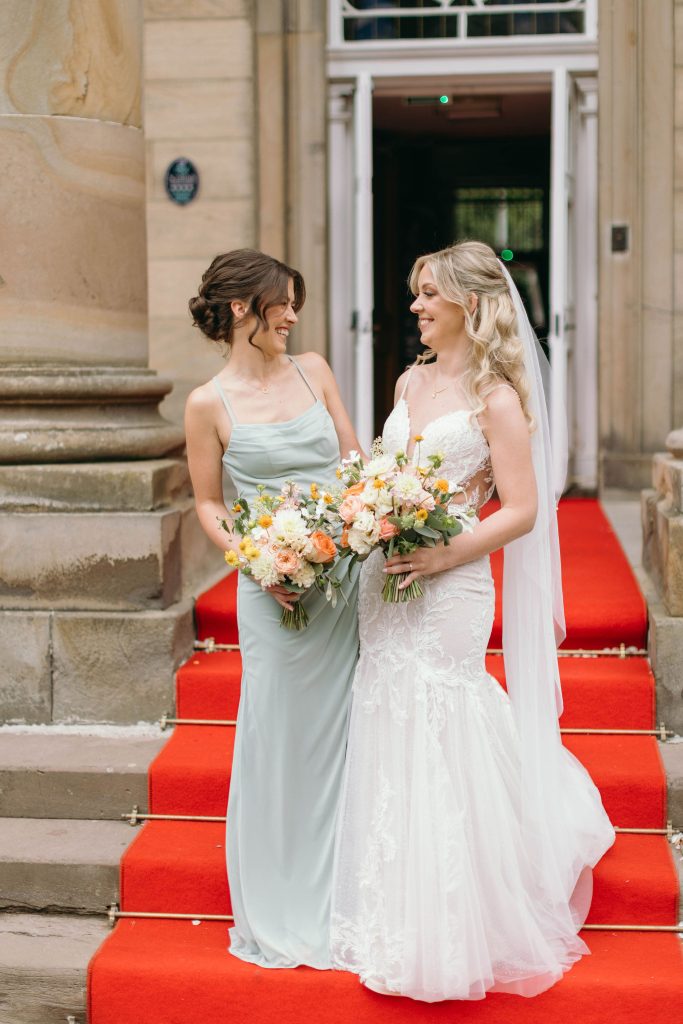 fraoch floral design bride in a white lace wedding dress with veil smiles at her bridesmaid while holding a bouquet on a red-carpeted steps outside a stone building.