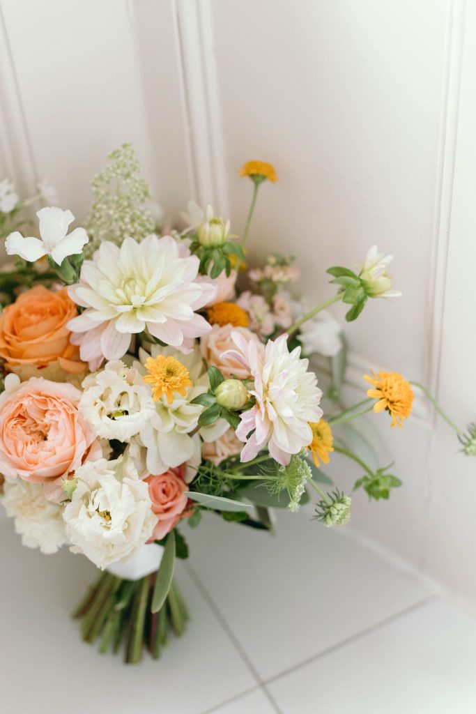 fraoch floral design bouquet of peach and white roses, pink dahlias, orange ranunculus, and yellow button mums against a light backdrop.