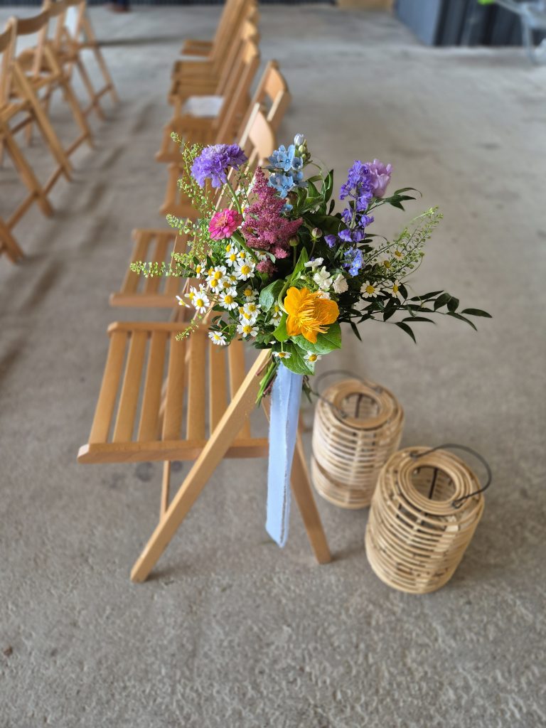 boreland-loch-tay-wedding-fraoch-floral-design 10 - fraoch floral design fraoch floral design colorful bouquet of wildflowers with daisies and purple blooms tied with a blue ribbon resting on a row of wooden chairs at a ceremony venue.