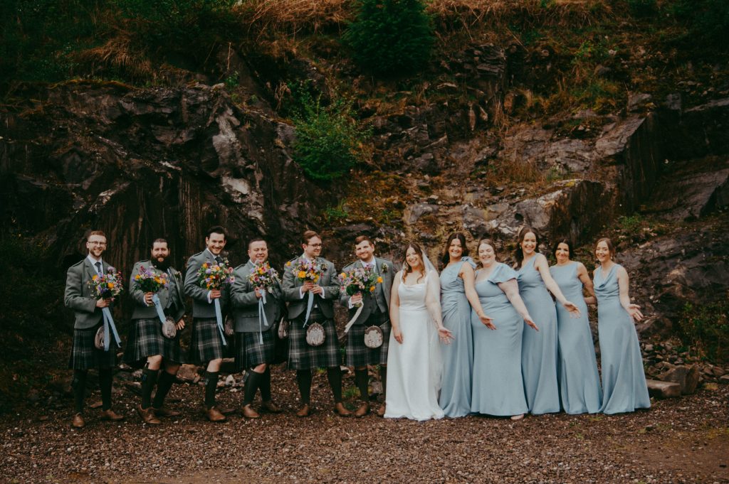 boreland-loch-tay-wedding-fraoch-floral-design 20 - fraoch floral design fraoch floral design wedding party posing outdoors: bridesmaids in light blue gowns and groomsmen in kilts hold bouquets, standing along a rocky hillside.