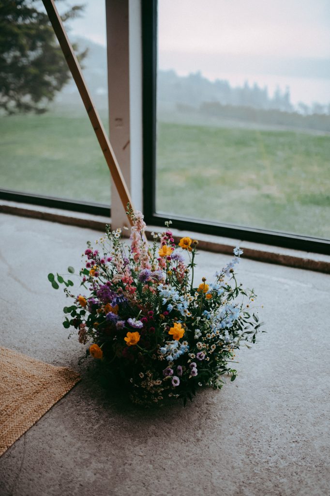 boreland-loch-tay-wedding-fraoch-floral-design 3 - fraoch floral design fraoch floral design bouquet of colorful wildflowers on a concrete floor by a window indoors, with a hazy green landscape outside.