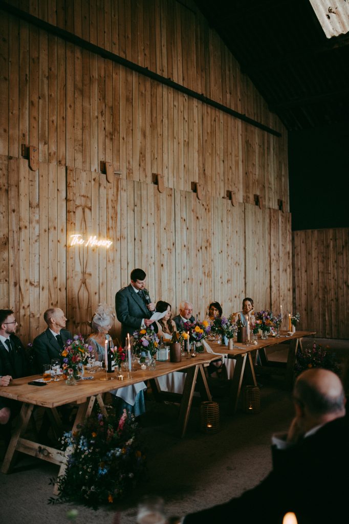 boreland-loch-tay-wedding-fraoch-floral-design 8 - fraoch floral design fraoch floral design rustic barn wedding scene with guests seated at a long wooden table, floral centerpieces, and candles; a man stands giving a speech.