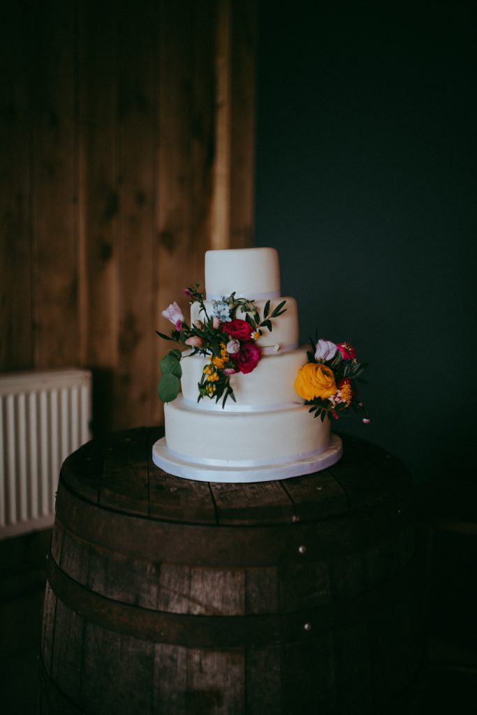 boreland-loch-tay-wedding-fraoch-floral-design 9 - fraoch floral design fraoch floral design three-tier white wedding cake decorated with colorful flowers on a rustic wooden barrel table