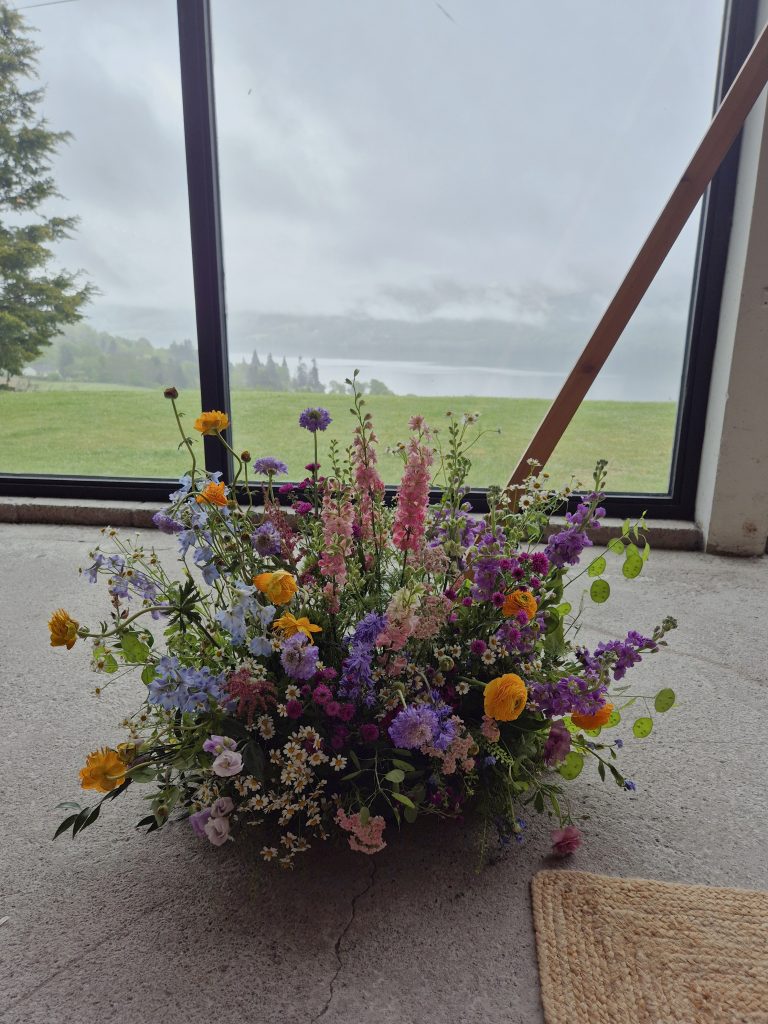 boreland-loch-tay-wedding-fraoch-floral-design 9 - fraoch floral design fraoch floral design a large colourful arrangement of wildflowers sits on a concrete floor by a slanted window, with a grassy landscape outside.