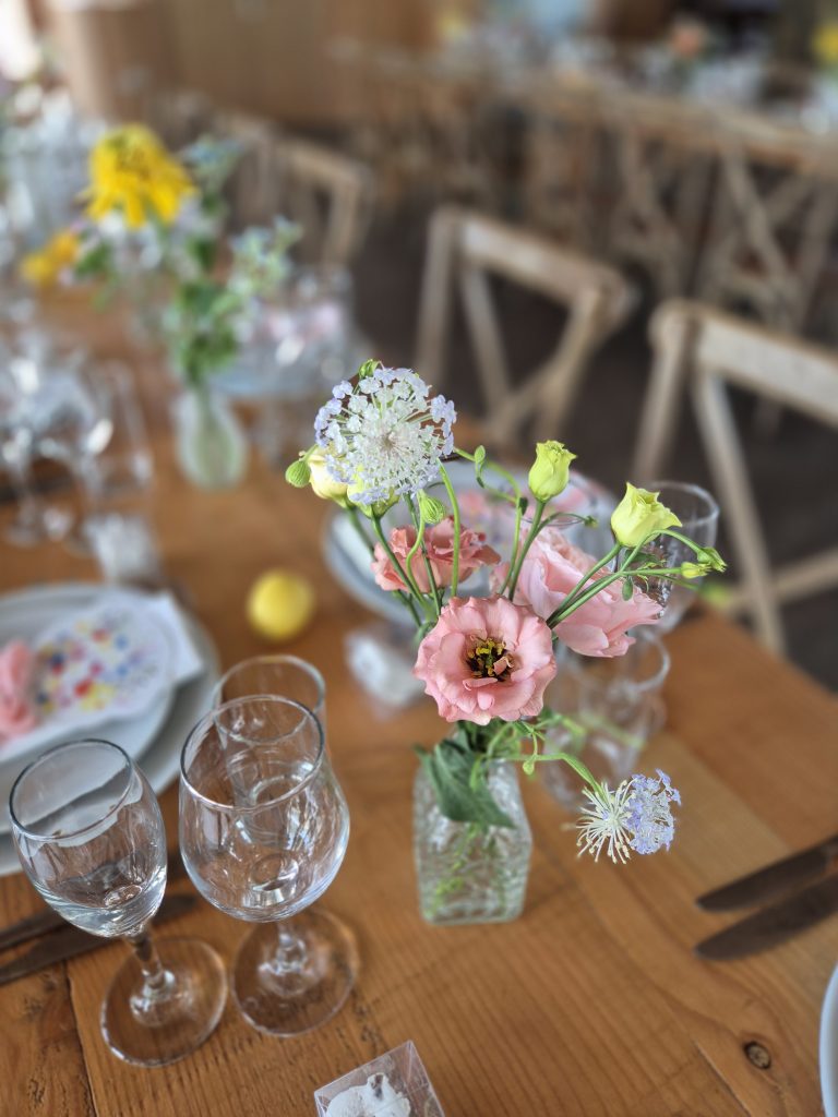 fraoch floral design close-up bouquet in a small glass vase on a wooden dining table with a blurred background of chairs and decor.