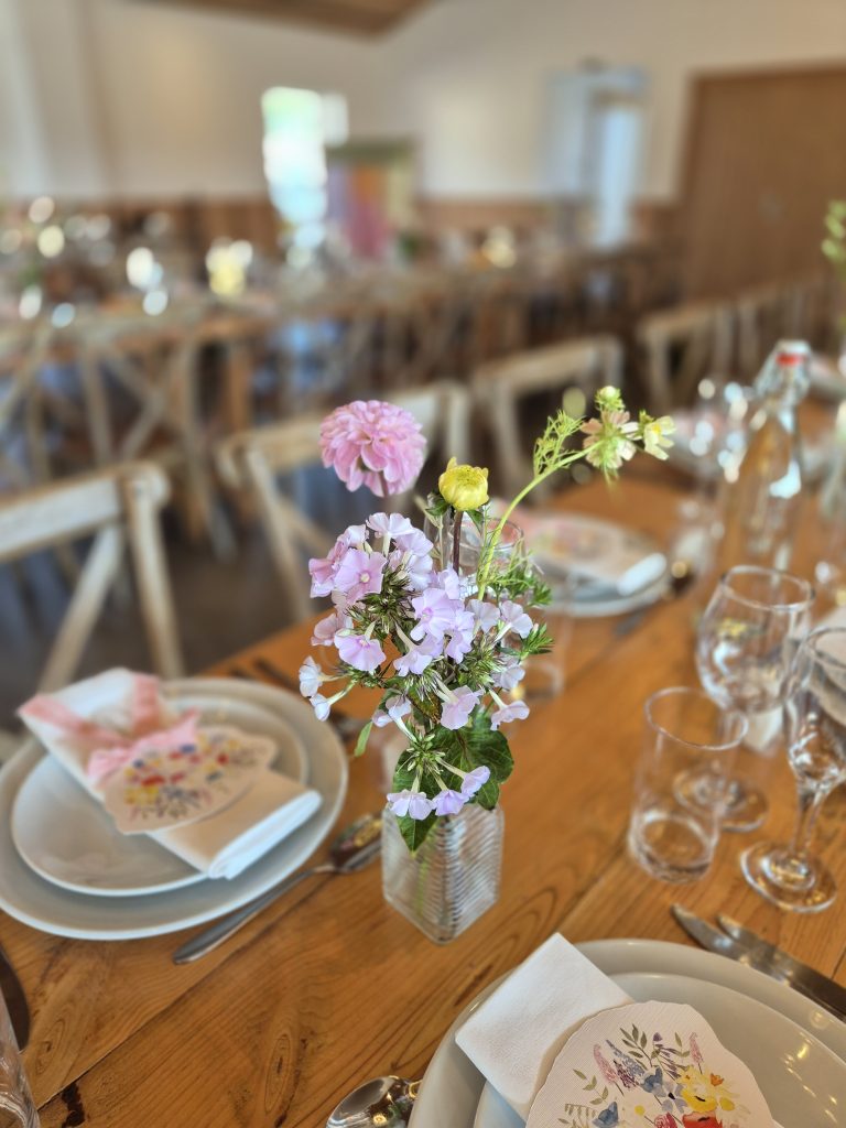 fraoch floral design pink and yellow flowers in a small glass vase on a wooden dining table, with plates, napkins, and glassware set for a meal in the foreground.