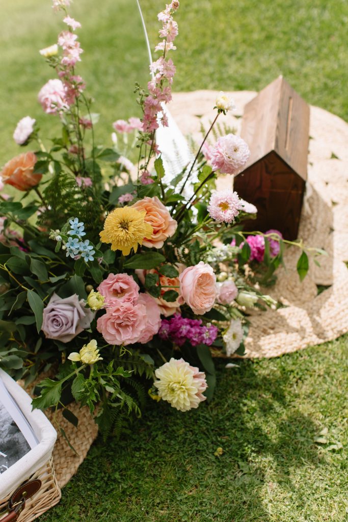 fraoch floral design colorful outdoor floral arrangement with pink, peach, and yellow blooms in a wicker basket on grass, with a small wooden birdhouse in the background