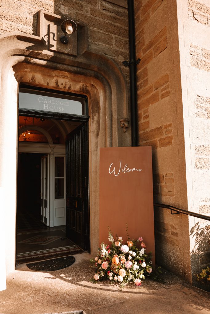 fraoch floral design entrance to carlogie house with an arched doorway, open doors, warm interior lighting, and a bouquet of pink and peach flowers outside.