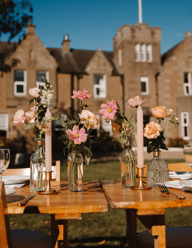 fraoch floral design outdoor dining table with glass bottles of pink flowers and tall pink candles, blurred historic building in background on a sunny day.