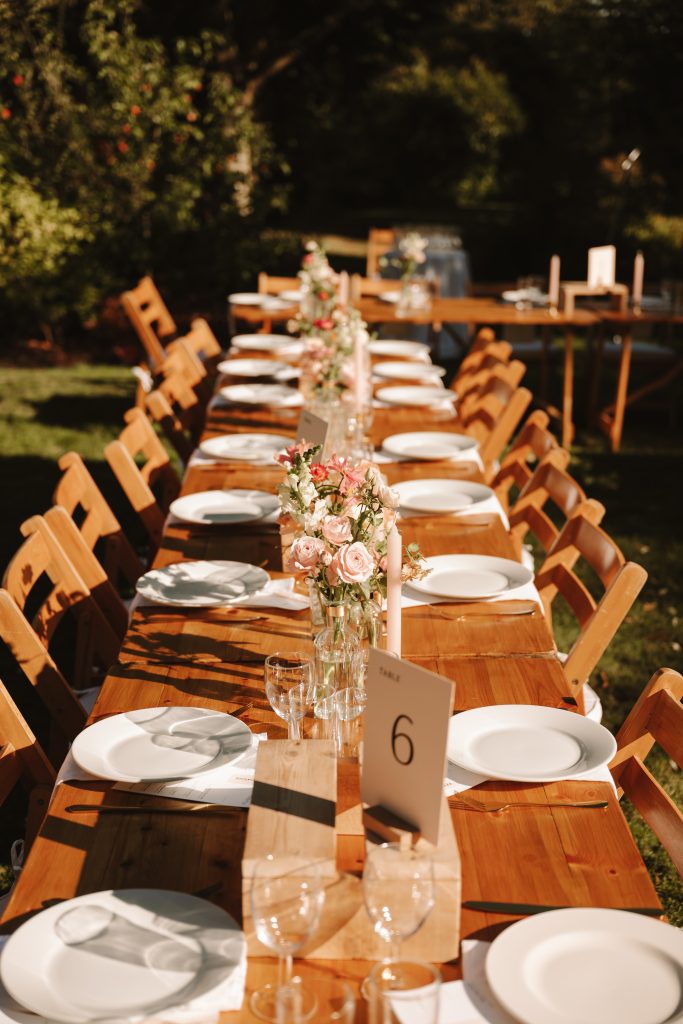 fraoch floral design long wooden outdoor dining table set for a garden party, with pink floral centerpieces, candles, and white plates.
