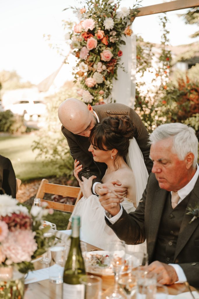 fraoch floral design bride with veil seated as a bald man hugs her from behind at an outdoor wedding under a pink floral arch.