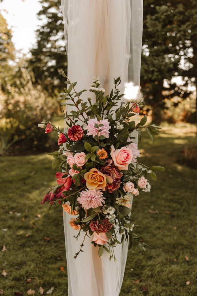 fraoch floral design floral arrangement of pink and peach roses and greenery tied to a sheer white fabric outdoors, likely a wedding display.