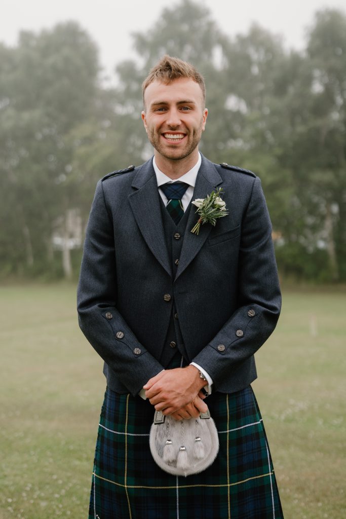 fraoch floral design smiling man in a dark suit with a boutonniere, wearing a plaid kilt, outdoors with trees in the background.