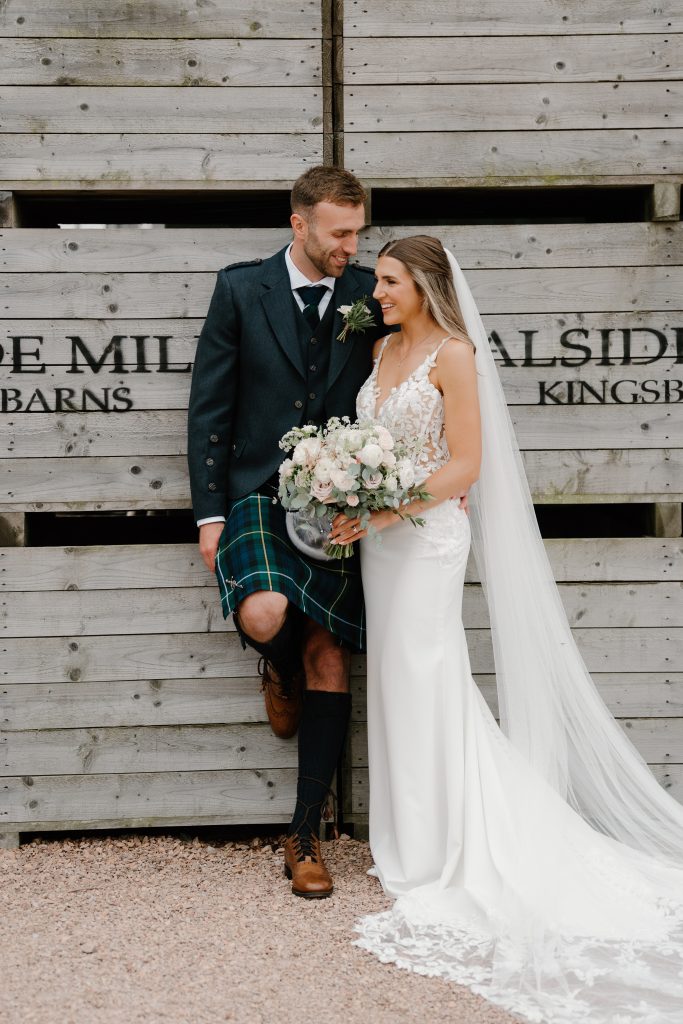 fraoch floral design bride and groom pose together against a weathered wooden wall; she holds a bouquet of white and blush flowers, he wears a dark formal jacket with a green tartan kilt and brown boots, smiling at each other under her veil.