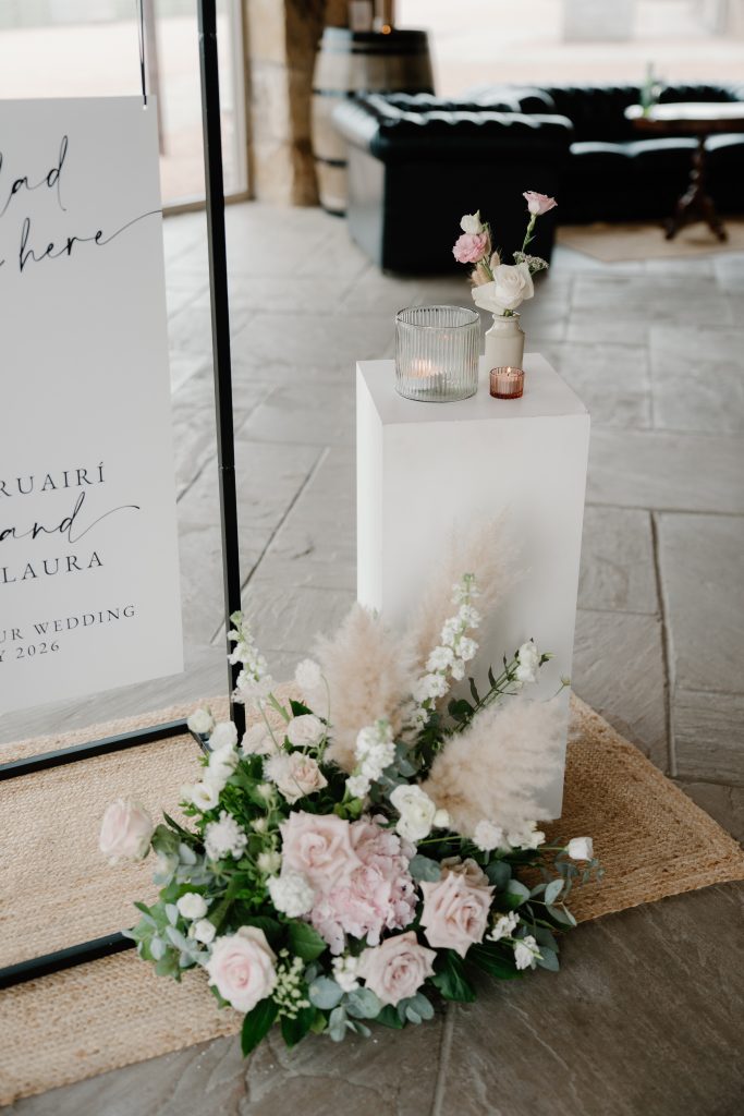 fraoch floral design wedding decor: white pedestal with a ribbed glass candle holder and a small pink vase, flanked by pastel flowers and pampas grass on a woven rug