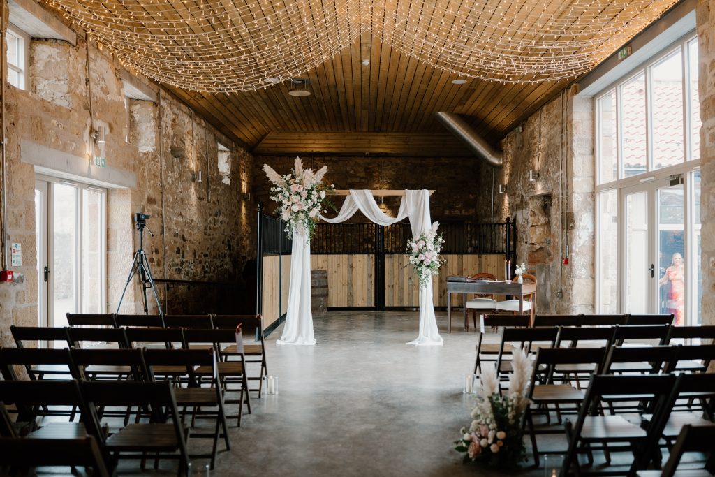 fraoch floral design rustic wedding ceremony setup in a stone-walled hall with a floral arch and white drapery, and rows of chairs.