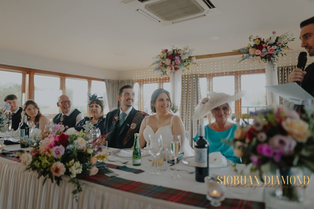 kerin kieran - fraoch floral design fraoch floral design bride in a white gown sits at a long, flower-filled wedding reception table as guests smile and chat beside her.