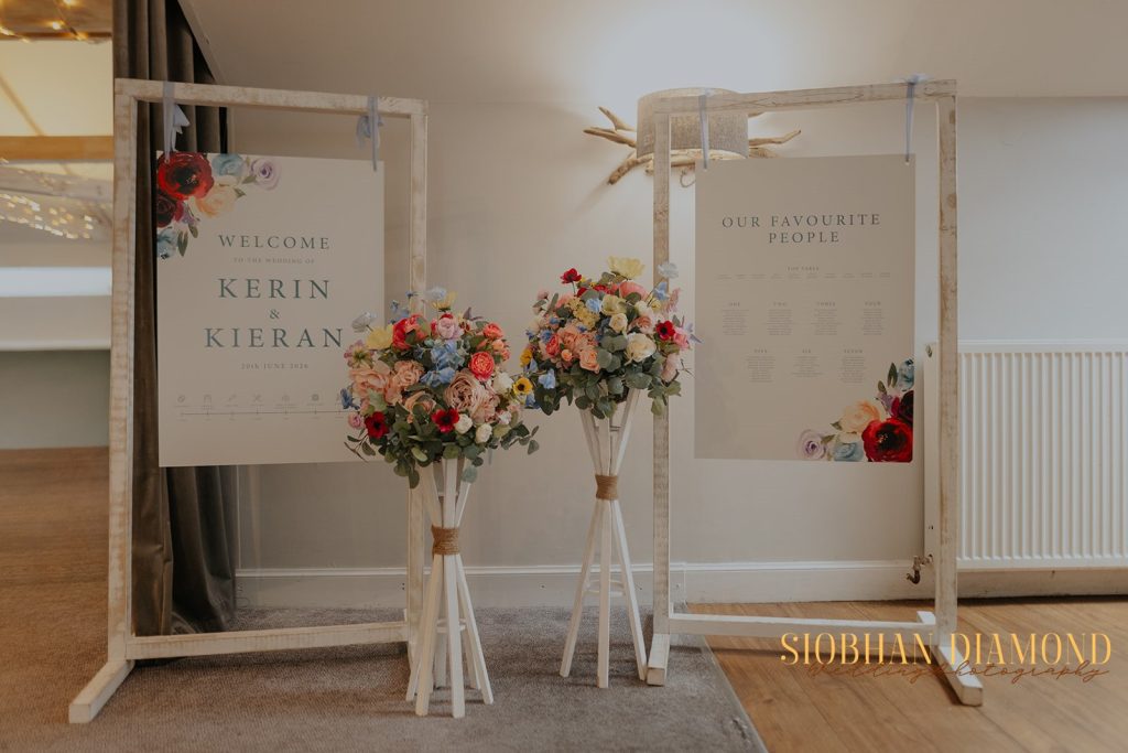 kerin kieran - fraoch floral design fraoch floral design wedding welcome sign reading 'welcome to the wedding of kerin & kieran' with floral borders on a wooden frame. two tall flower stands flank it.