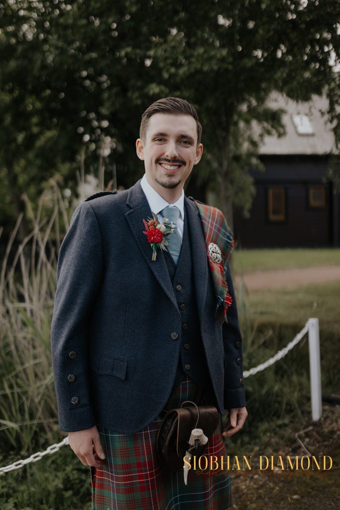 kerin kieran - fraoch floral design fraoch floral design smiling man in a dark blue suit and tartan kilt with a red boutonniere, standing outdoors in a garden setting.