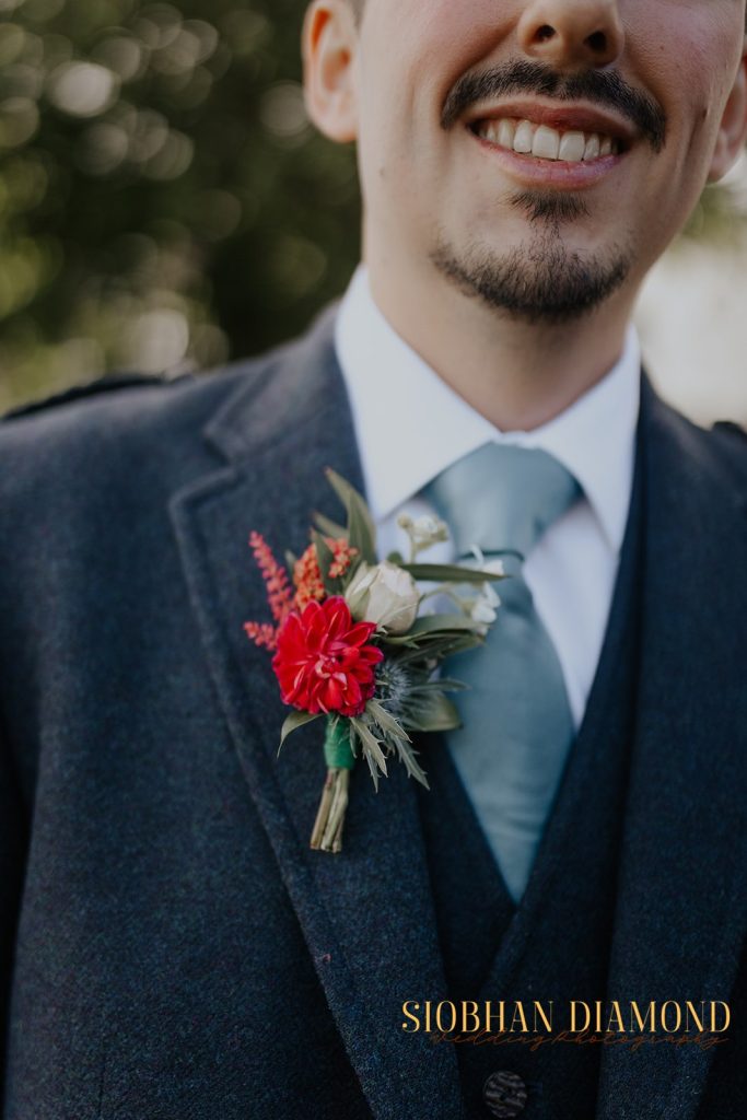 kerin kieran - fraoch floral design fraoch floral design close-up of a groom in a dark suit with a teal tie, smiling, wearing a red boutonniere on his lapel.