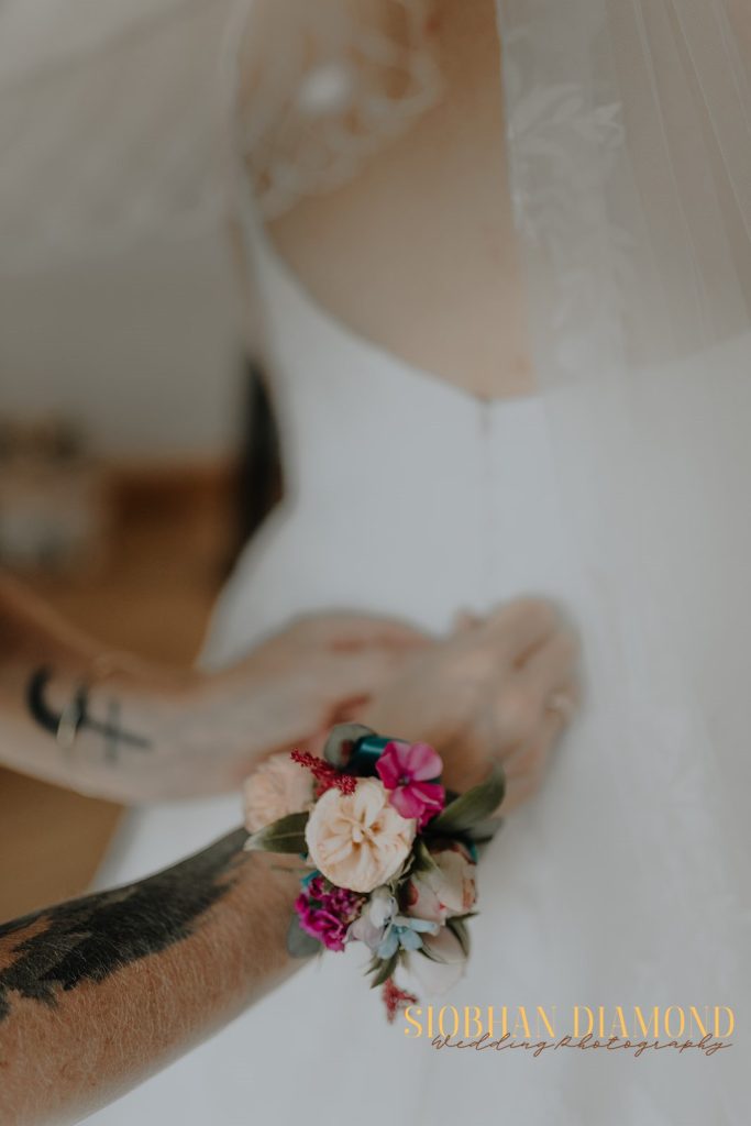 kerin kieran - fraoch floral design fraoch floral design close-up of a tattooed arm adjusting a colorful wedding boutonniere on the bride's dress with veil in the background, soft focus.