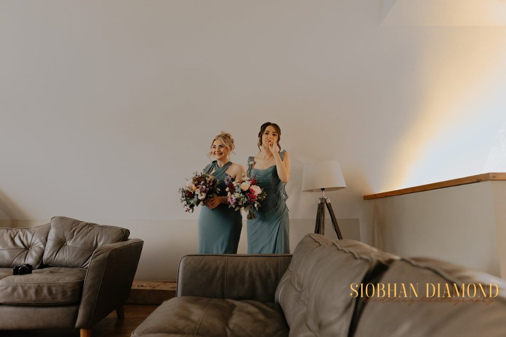 kerin kieran - fraoch floral design fraoch floral design two bridesmaids in teal dresses hold vibrant bouquets and pose indoors beside a gray couch and lamp in a bright room.