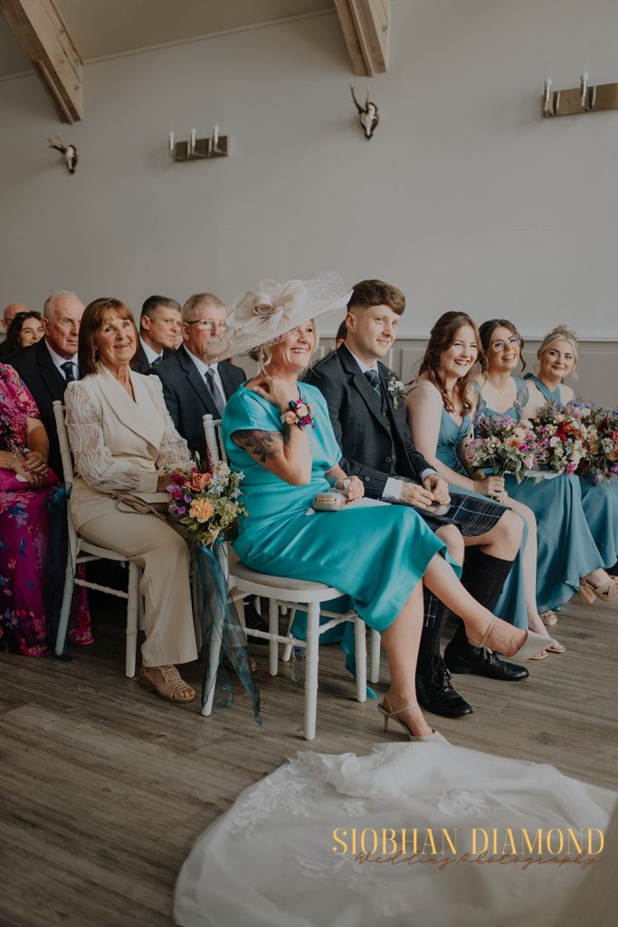 kerin kieran - fraoch floral design fraoch floral design wedding guests seated during a ceremony, including a woman in a teal dress with a large fascinator and a man in a suit, smiling with bouquets nearby.