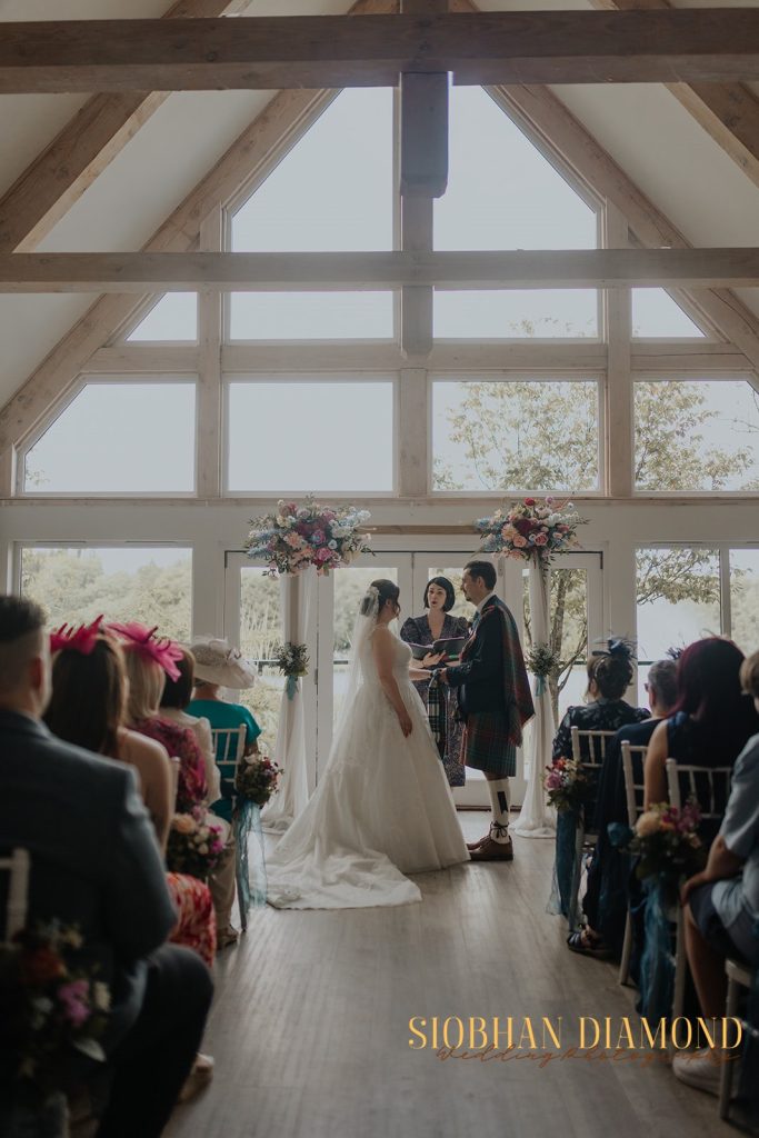 kerin kieran - fraoch floral design fraoch floral design bride and groom face each other with an officiant in a bright, wooden-beam ceremony space, guests seated around them.