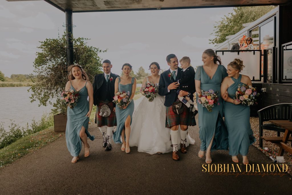 kerin kieran - fraoch floral design fraoch floral design bride and wedding party walking along a lakeside path, bridesmaids in teal dresses with bouquets, groomsmen in kilts and vests, smiling together on wedding day.