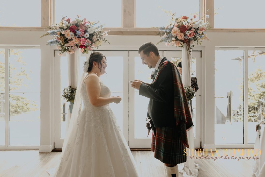 kerin kieran - fraoch floral design fraoch floral design bride and groom exchange rings during a sunny wedding indoors, with large floral arches behind them and a lace wedding gown on the bride.