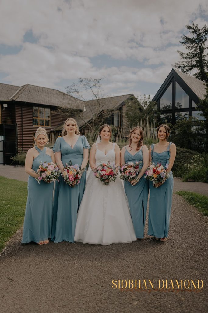 kerin kieran - fraoch floral design fraoch floral design bride in a white gown stands with four bridesmaids in blue dresses outdoors, each holding colorful bouquets, in front of a modern building.