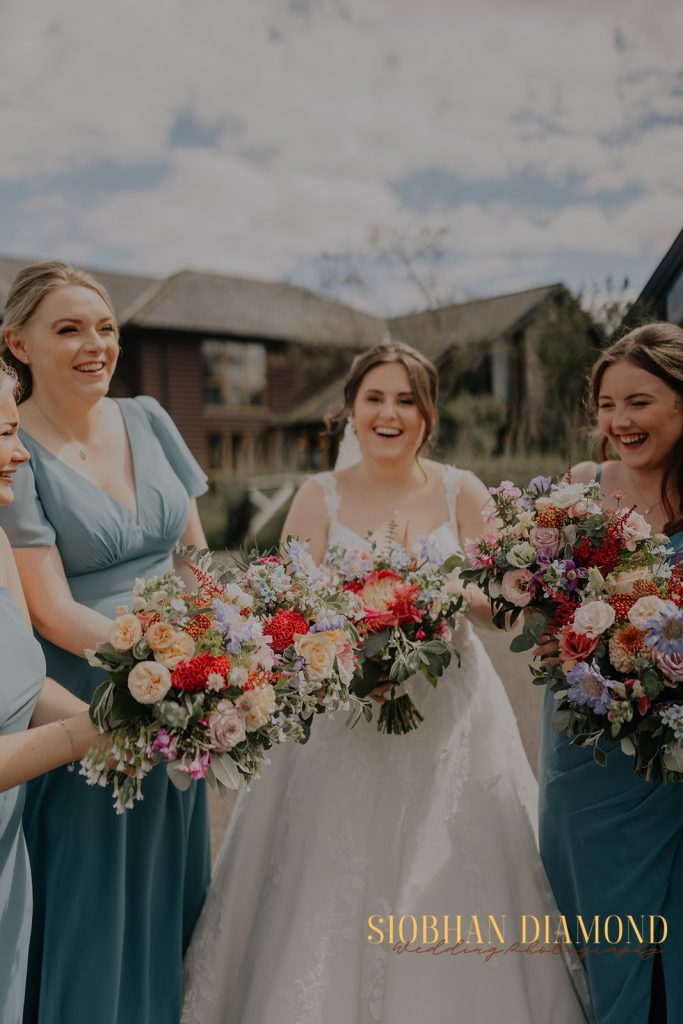 kerin kieran - fraoch floral design fraoch floral design bride in a white gown surrounded by her bridesmaids in blue dresses, all smiling while holding vibrant bouquet arrangements outdoors.