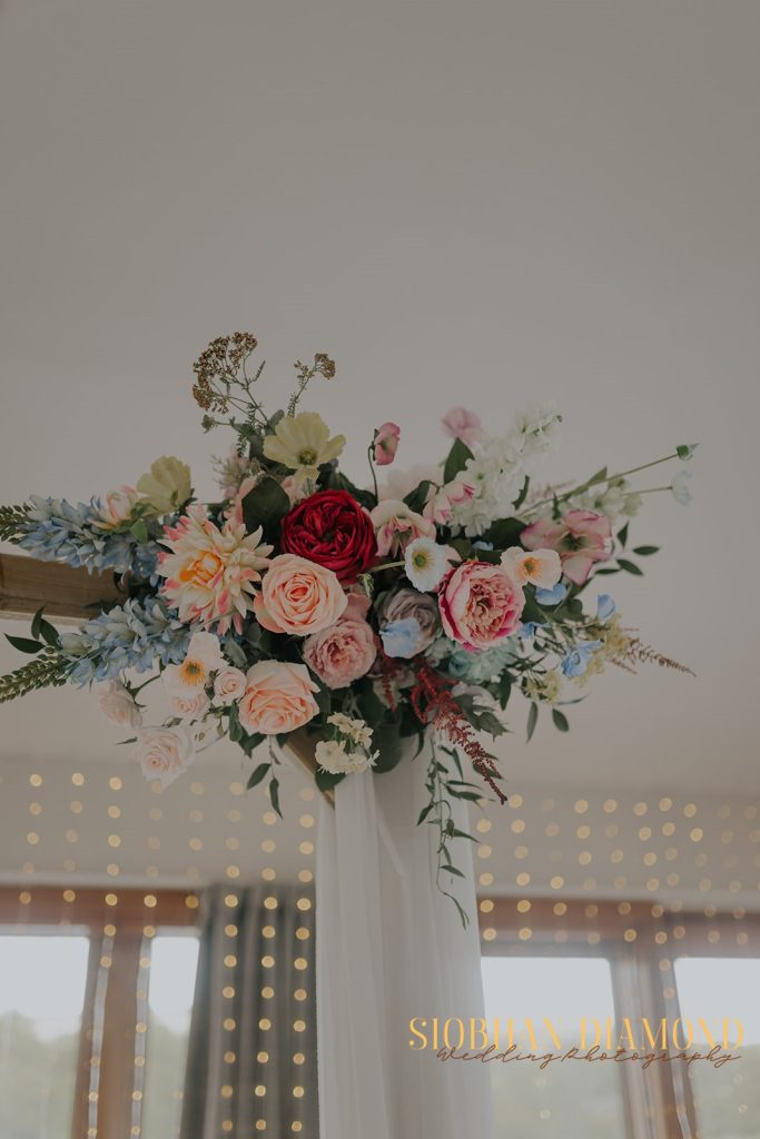 kerin kieran - fraoch floral design fraoch floral design colorful wedding bouquet with pink, peach, and blue flowers atop a white draped stand, warm lights in the background.