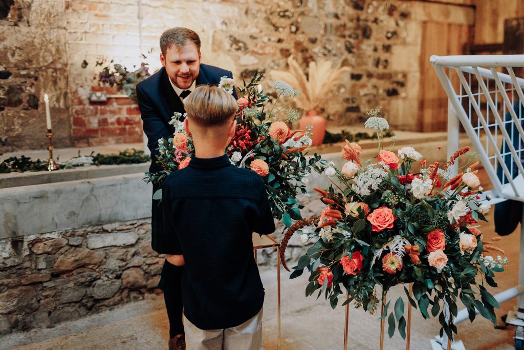 fraoch floral design a man in a suit stands behind a boy admiring a large colorful floral arrangement at a rustic indoor venue.