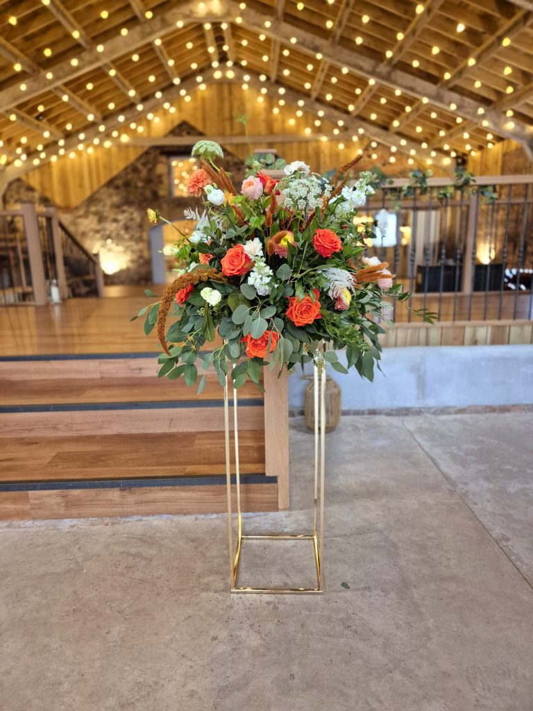 fraoch floral design tall floral centerpiece with orange roses, white blooms, and greenery on a gold pedestal in a rustic, vaulted venue with warm string lights above it.