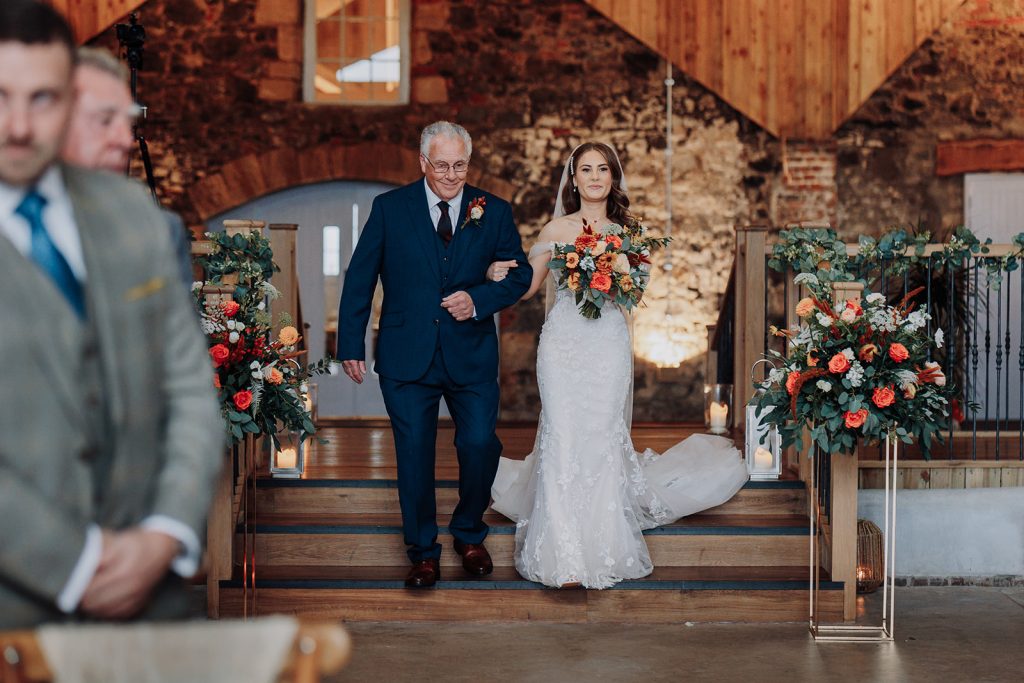 fraoch floral design bride in a lace wedding gown, veil, and holding a bright bouquet walks down the wooden aisle arm-in-arm with an older man in a navy suit at a rustic venue.