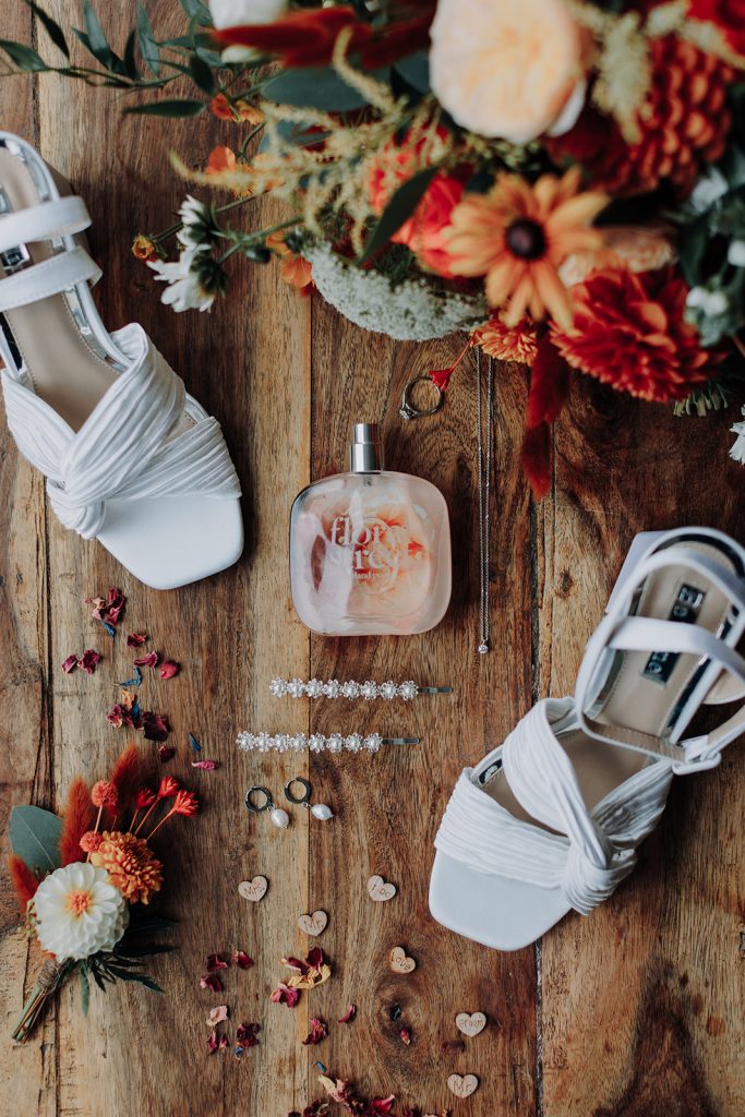 fraoch floral design white wedding sandals beside a peach-and-orange bouquet on a wooden floor, with perfume bottle and scattered petals nearby.