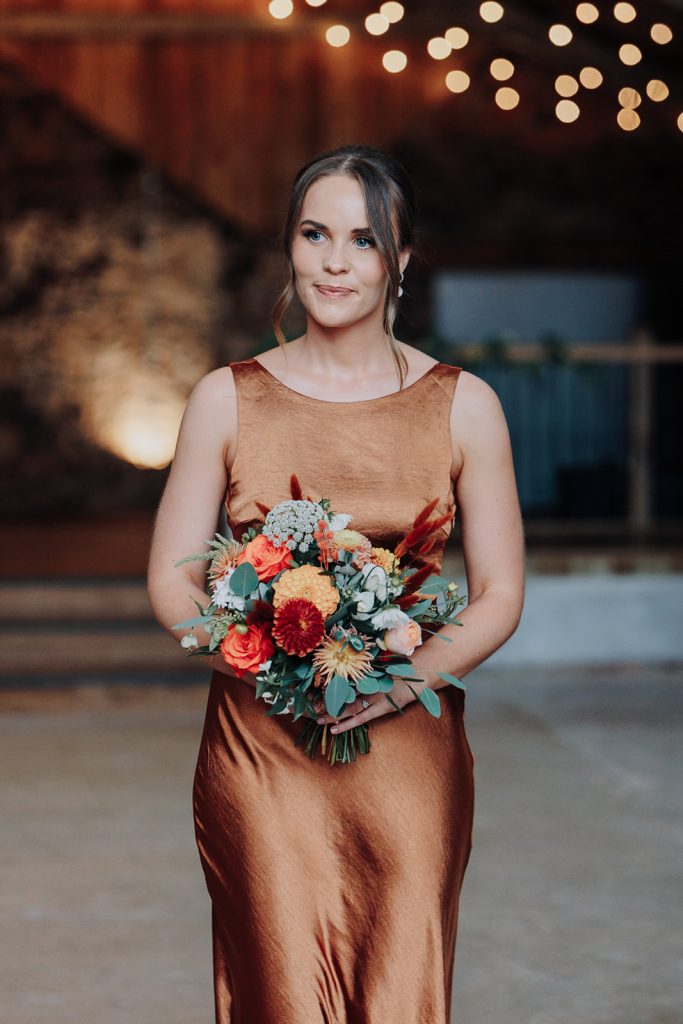 fraoch floral design woman in a copper satin dress holding a bright bouquet of orange and cream flowers, outdoors with warm string lights in the background.