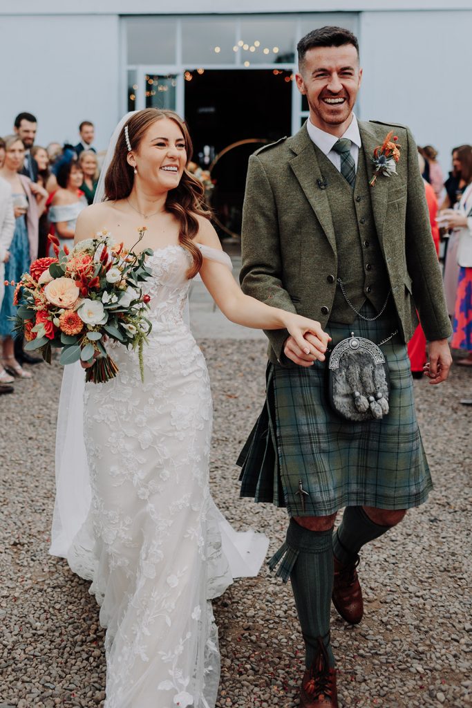 fraoch floral design bride in a lace wedding gown holding a vibrant bouquet, smiling, walking hand in hand with a groom in a green plaid kilt outside a venue with guests in the background.