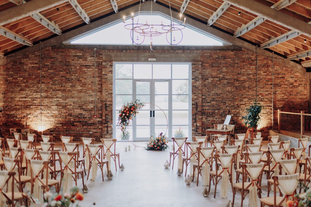 fraoch floral design wedding ceremony setup in an industrial loft with brick walls, wooden beams, and white chairs facing a circular floral arch near glass doors.