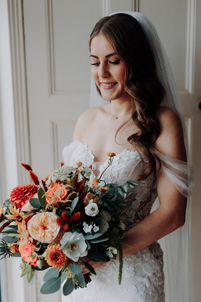 fraoch floral design bride in a white lace wedding gown holding a colorful bouquet near her waist, veil flowing behind her, smiling softly by a doorway.