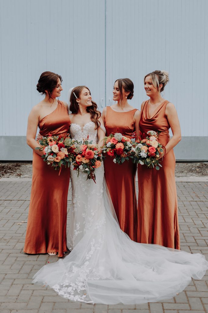 fraoch floral design bride in a white lace wedding gown stands with three bridesmaids in copper satin dresses, all holding bouquets, in front of a pale blue wall.